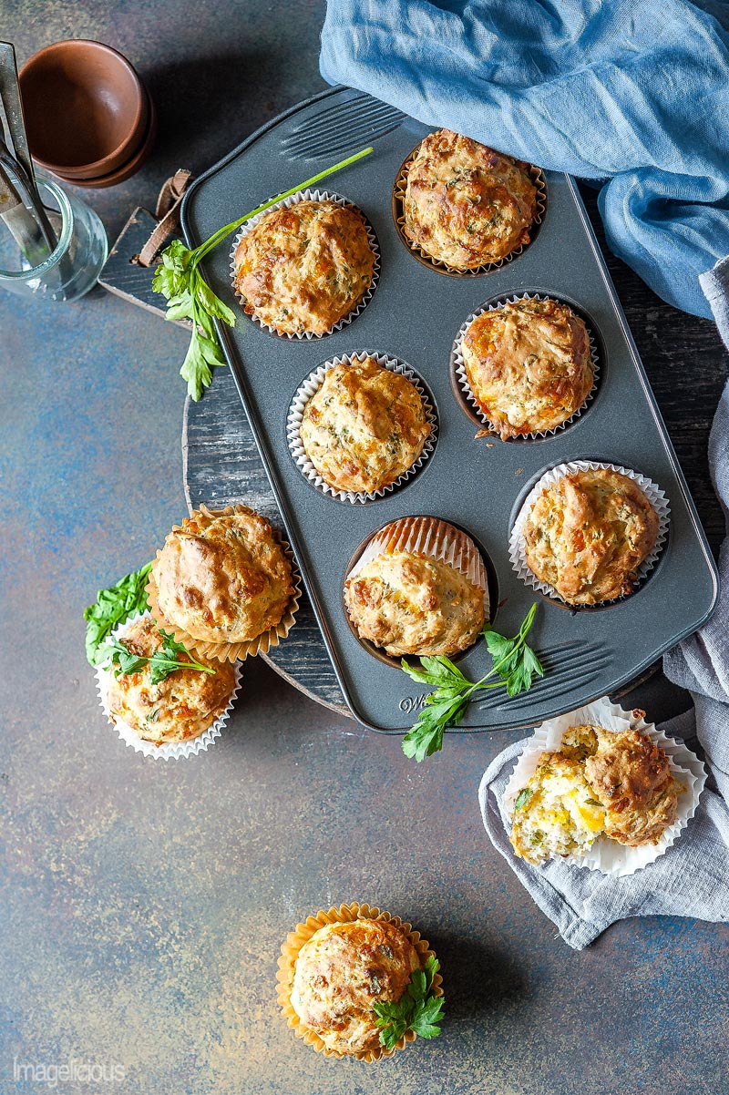 Top down view of a pan of Herbs and Cheese Muffins with a few more muffins around the pan. Some sprigs of parsley around and a few forks and spoons and a napkin