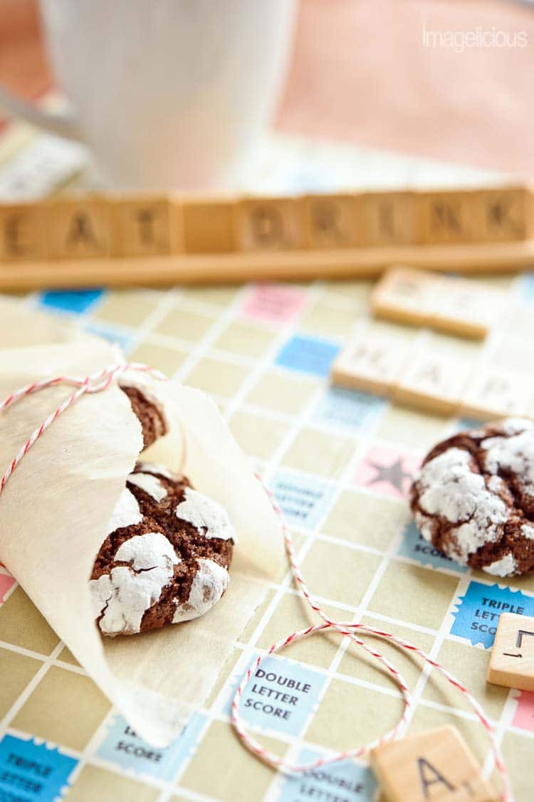 Another close up of Chocolate-Mint Crackle Cookies, wrapped in parchment paper with festive red string. All on a scrabble board with words "eat drink" blurry in the background