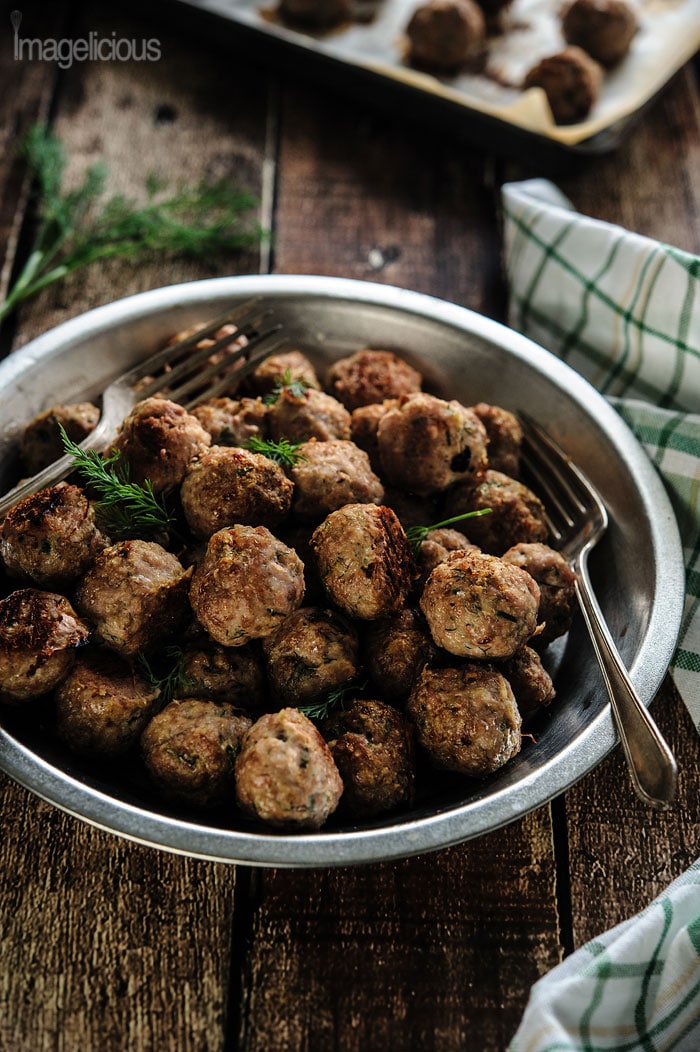 A big plate of Dill Meatballs with a couple of forks. A green napkin to the right and a sheet pan with more meatballs in the background.