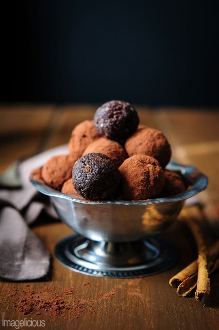 A small metal bowl filled with truffles on a wooden table with cinnamon sticks to the right. Most truffles are rolled in cocoa powder but two of them are not rolled and are darker in colour