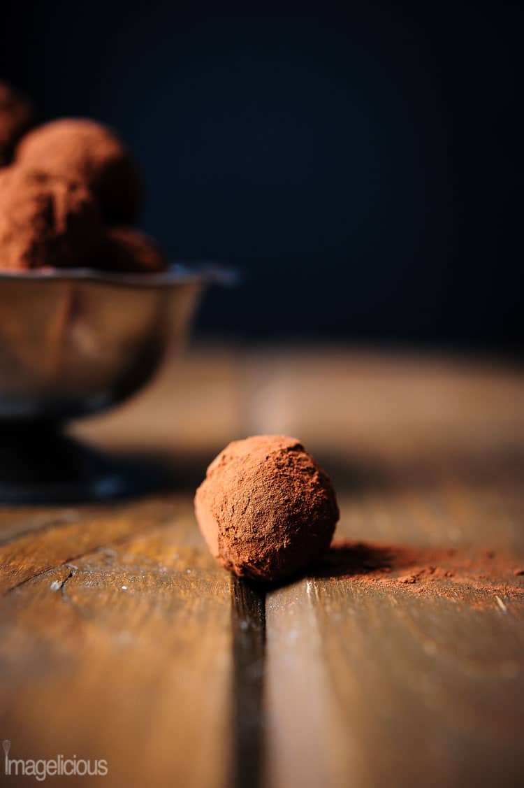 Close up of a cocoa rolled truffle on a wooden table. A little bit of the bowl with truffles is in the background, blurred