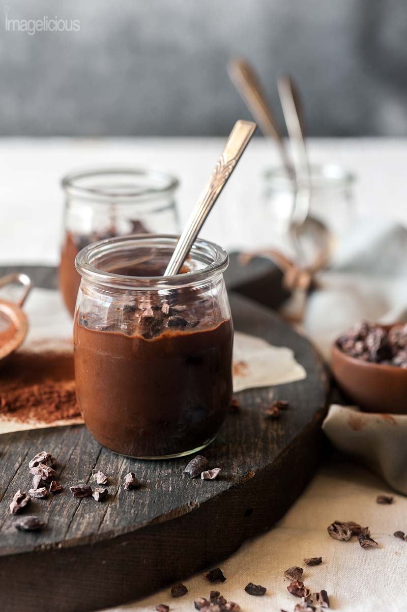 Small jars filled with Avocado Chocolate Pudding on a wooden cutting board. A few cocoa nibs are scattered around the jars and a dusting of cocoa powder