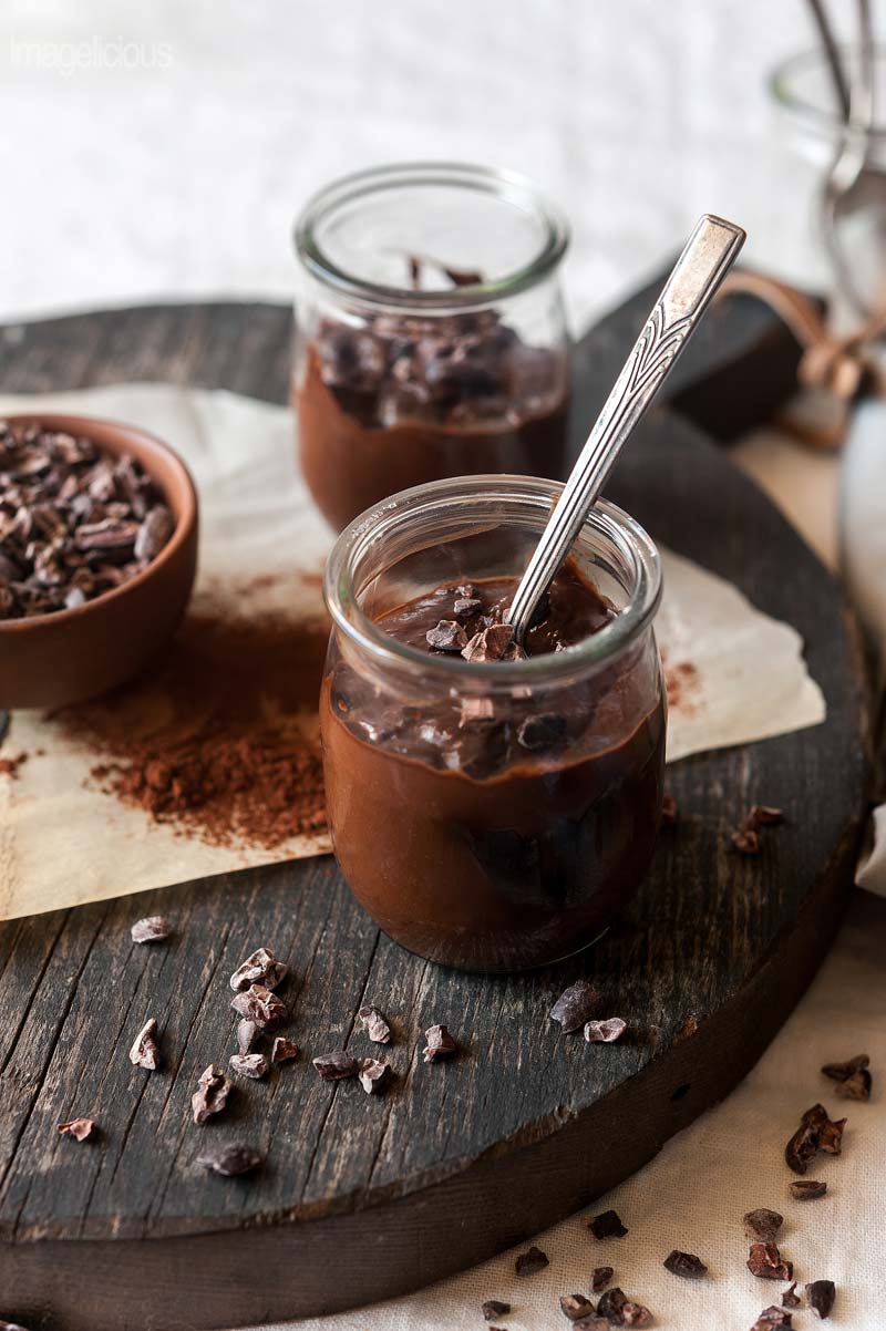 Two jars of Avocado Chocolate Pudding on a cutting board with a small bowl of cocoa nibs