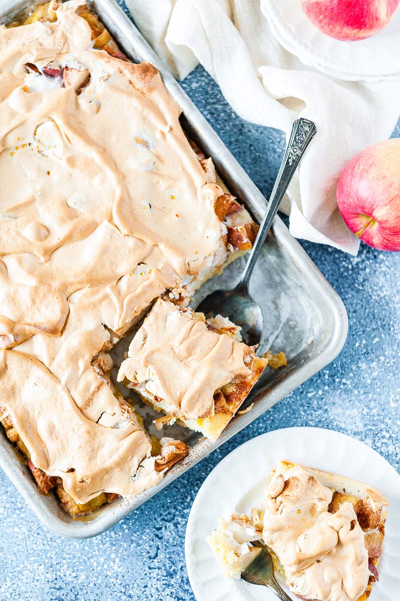 Top down view of the sheet pan with Apple Meringue Tart, one slice is cut and is resting on a cake server inside the pan. A plate with another slice and a fork at the bottom of the picture. A couple of apples are positioned around the sheet pan on a napkin and plates
