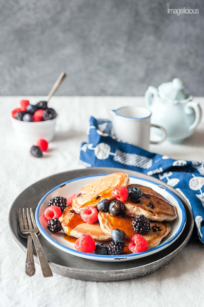 A plate of bumbleberry pancakes with a sprinkling of fresh berries on top, a bowl of berries in the background, and a jar with maple syrup, blue napkin and fork next to the plate.