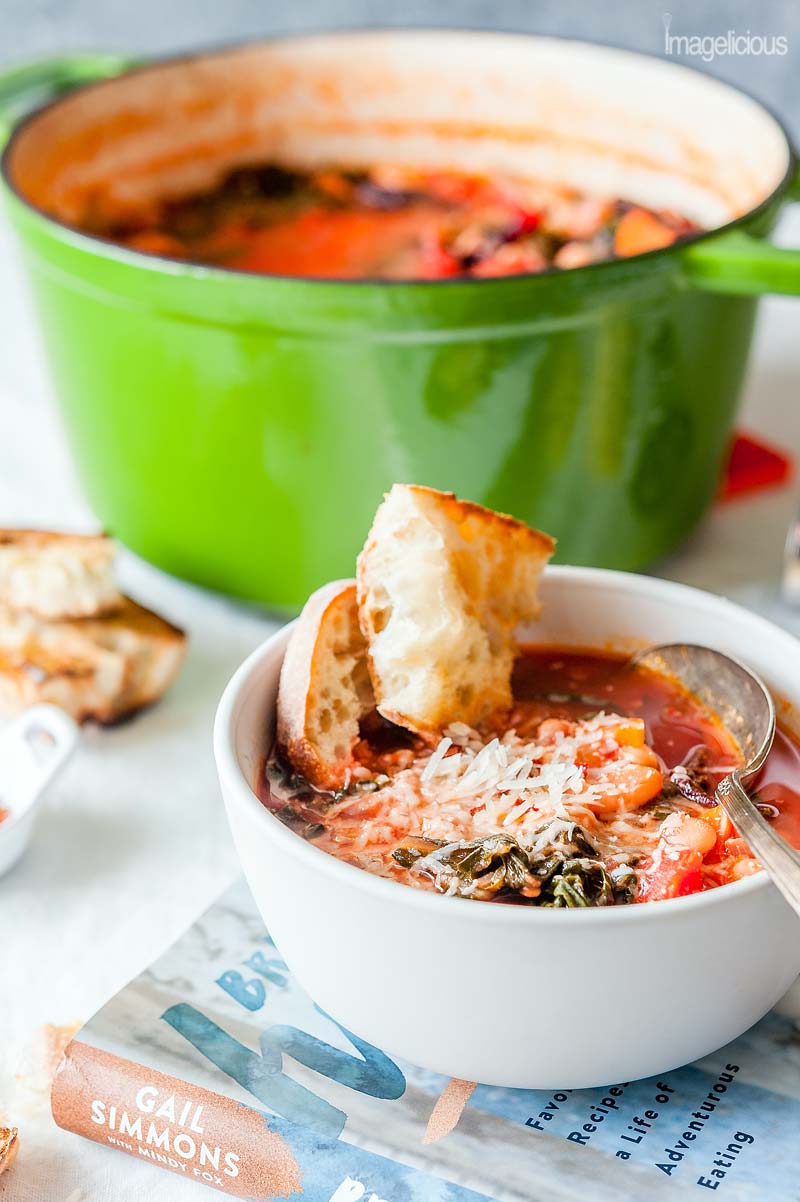 A bowl of vegetable soup called Ribollita with a few pieces of bread in it. Some bread and cheese is visible in the background. A big green pot of soup is behind the bowl. The bowl is standing on top of a cookbook called Bringing it Home