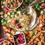 Sheet pan with cooked thanksgiving dinner. Turkey breast in the center of the pan with a few slices already cut, roasted brussels sprouts and sweet potatoes around the turkey. Two ramekins with cooked stuffing and a small ramekin with cranberry sauce. Parsley is sprinkled over everything. A knife and a fork are next to turkey