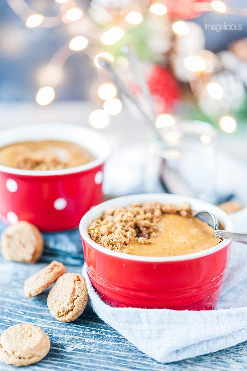 Close up of the red ramekins with gingerbread cheesecake and ginger snap cookies scattered around. Festive christmas lights are in the background, blurry