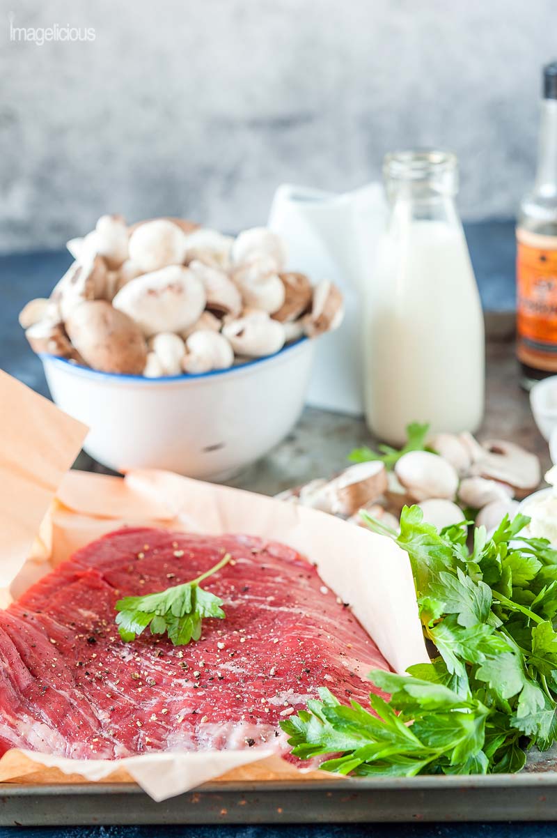 All the raw ingredients to make family-style steak with mixed mushrooms. On a tray there's raw steak, a bowl with mushrooms, parsley, butter, a bottle of milk