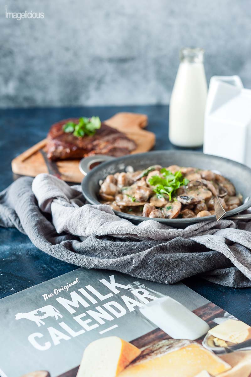 Canadian milk calendar is in the forefront of the picture, behind it there's a pan with creamy mushrooms, behind it a cutting board with the cooked steak, behind is a bottle with milk