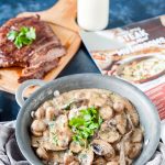 A pan with creamy mushrooms, a cutting board with the steak behind it, a calendar that's open to the page displaying this same recipe, a bottle of milk behind the calendar