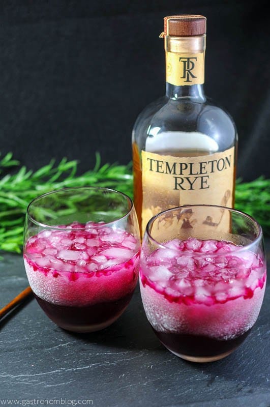 Two frosty glasses filled with pink beet cocktail and a bottle of rye behind