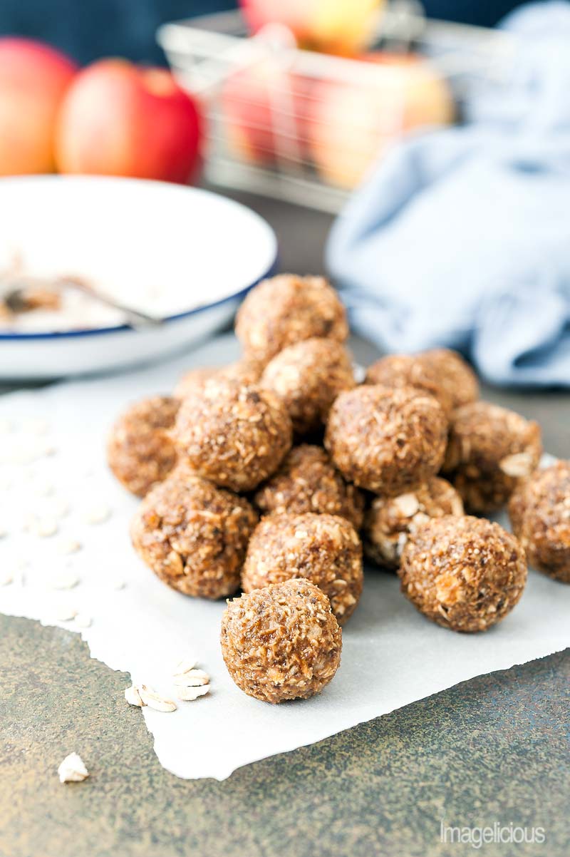 A pile of Apple Pie Energy Balls on a parchment paper with a few oats scattered around. A napkin, plate, and a few apples are in the background