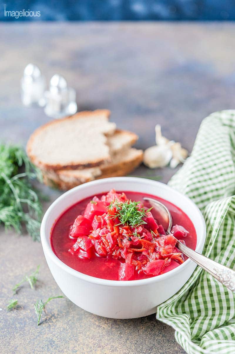 White bowl of borscht with a spoon. Green napkin to the right, a few slices of rye bread in the background. A few sprigs of dill in the background as well