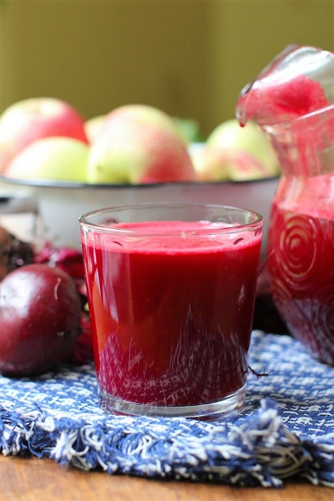 Close up of a glass filled with apple beet juice. A bowl of apples and a pitcher of juice are in the background