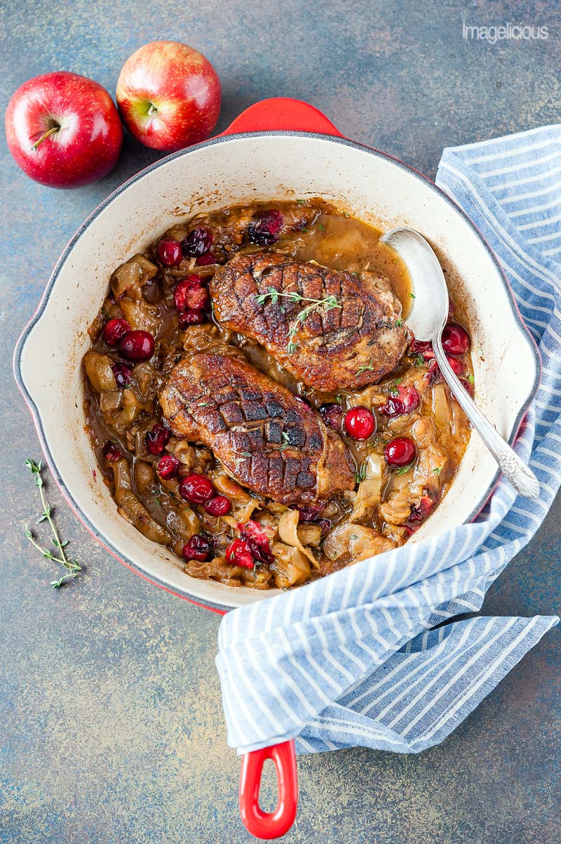 Top down view of a red pan filled with Two Braised Duck Breasts with Apples and Onions, a few cranberries are visible in the sauce. A few fresh thyme leaves are sprinkled throughout. Two whole ontario apples are next to the pan.