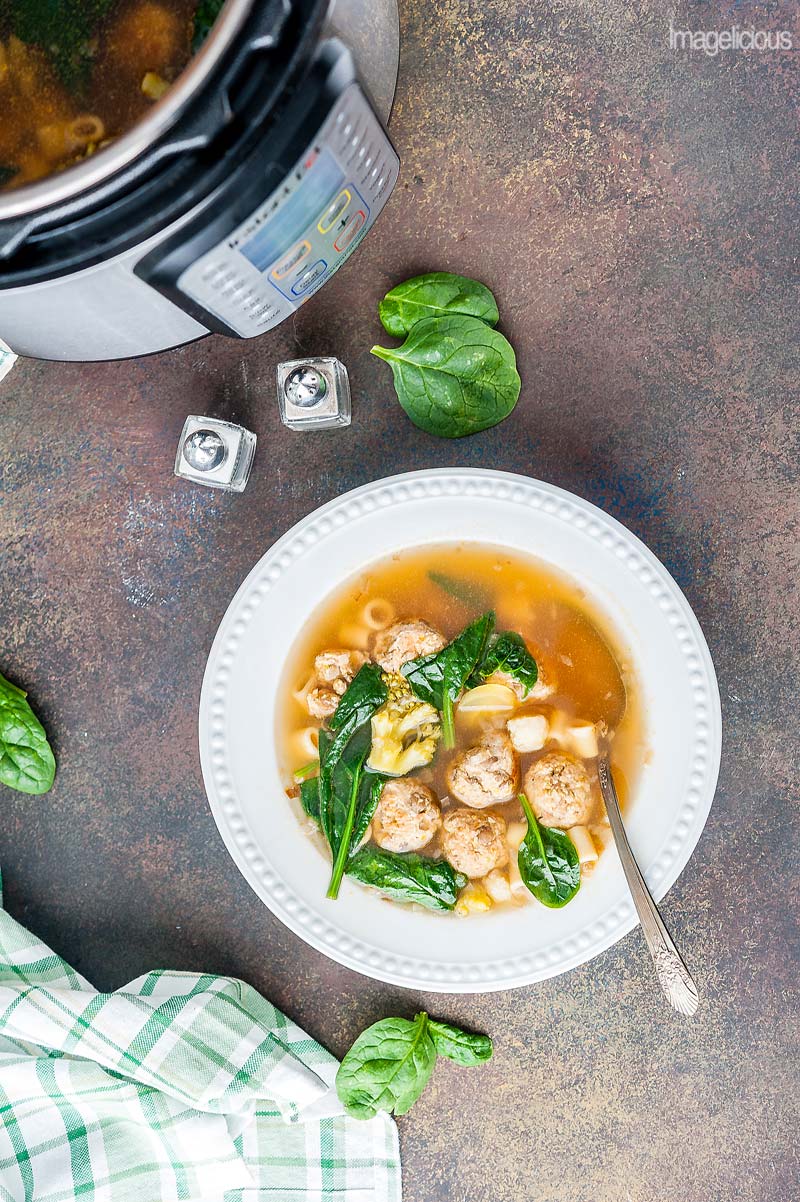 Top down view of a plate filled with Instant Pot Meatball soup with a spoon. A few spinach leaves are around the plate. Instant Pot is also visible in the corner