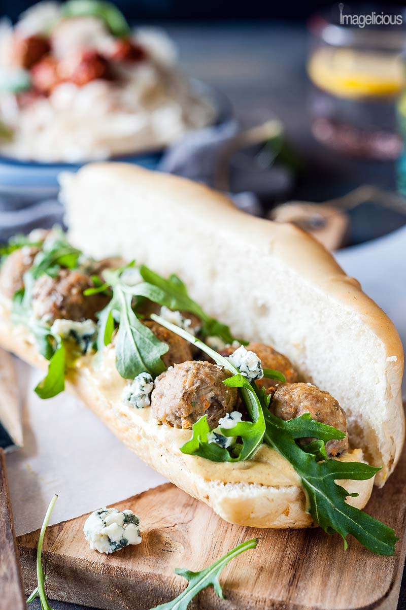 Closeup of a long bread roll split in half, spread with mustard, and filled with Lentil-Turkey Meatballs, blue cheese and arugula. More food and drink is visible in the background, blurry