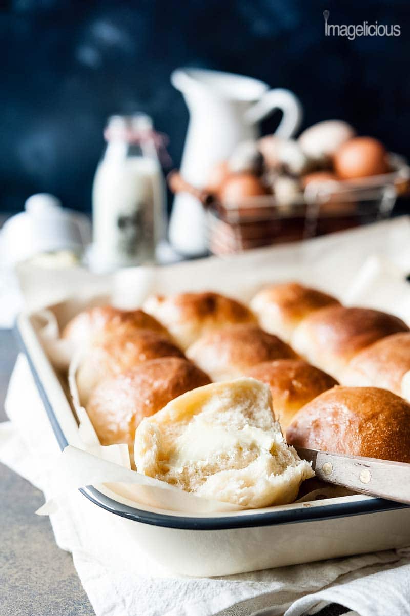 Closeup view of a pan of Instant Pot No Knead Dinner Rolls with one of the rolls open and buttered. In the background there's a basket of eggs, white pitcher, and a bottle of milk, all blurred