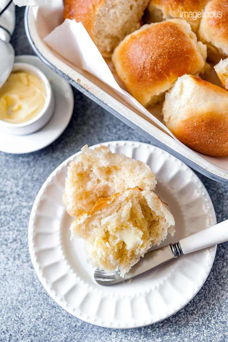 Top down view of a plate with an Instant Pot No Knead Dinner Roll that is torn in half and buttered. Above the plate a pan with more dinner rolls is visible and a small dish with butter