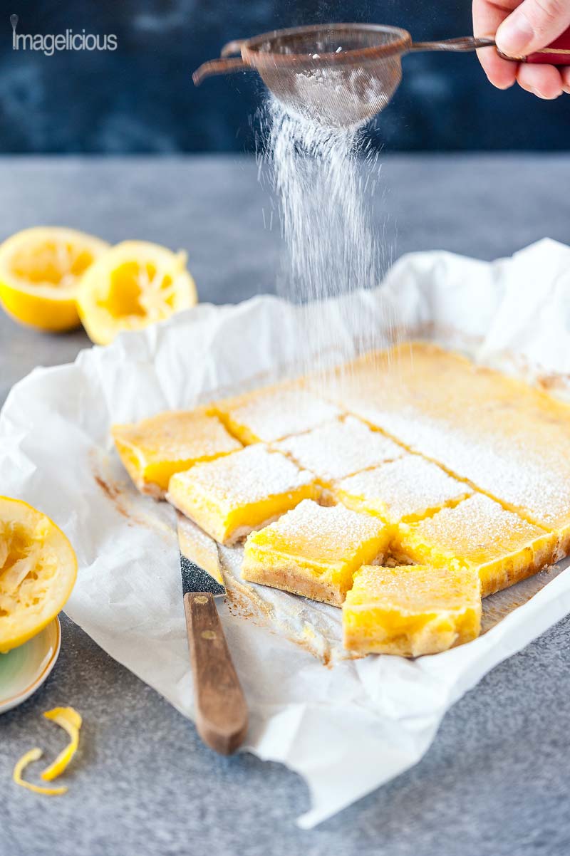 Side view of Coconut Lemon Bars with a hand adding icing sugar with a small sieve. 