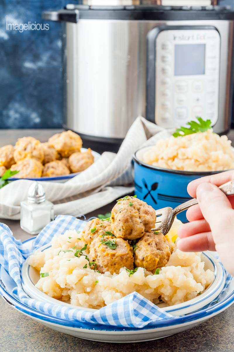 Closeup view of a plate filled with Instant Pot Meatballs and Mashed Potatoes and a hand is holding a fork and picking up one of the meatballs. In the background another two bowls are visible filled with mashed potatoes and meatballs. An Instant Pot is in the background, blurred.