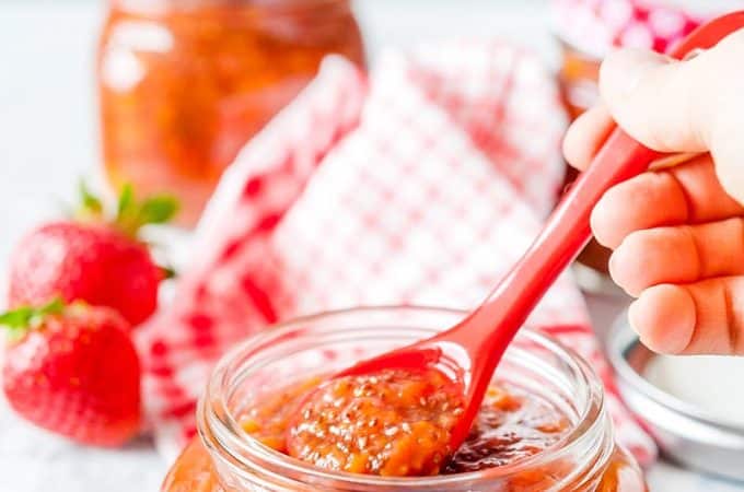 Closeup of a Strawberry Chia Jam with a hand holding a spoon inside a jar. Fresh strawberries in the background with a red napkin and another jar