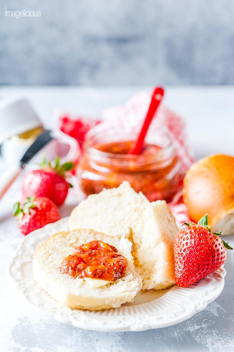 Small plate with a soft dinner roll spread with butter and Strawberry Chia Jam. A jar of the jam in the background with fresh strawberries around it