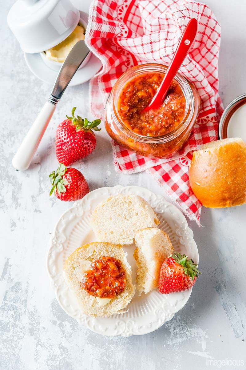 Top down view of a small plate with a dinner roll spread with the Strawberry Chia Jam. An open jar of jam is next to the plate, a few fresh strawberries are around it