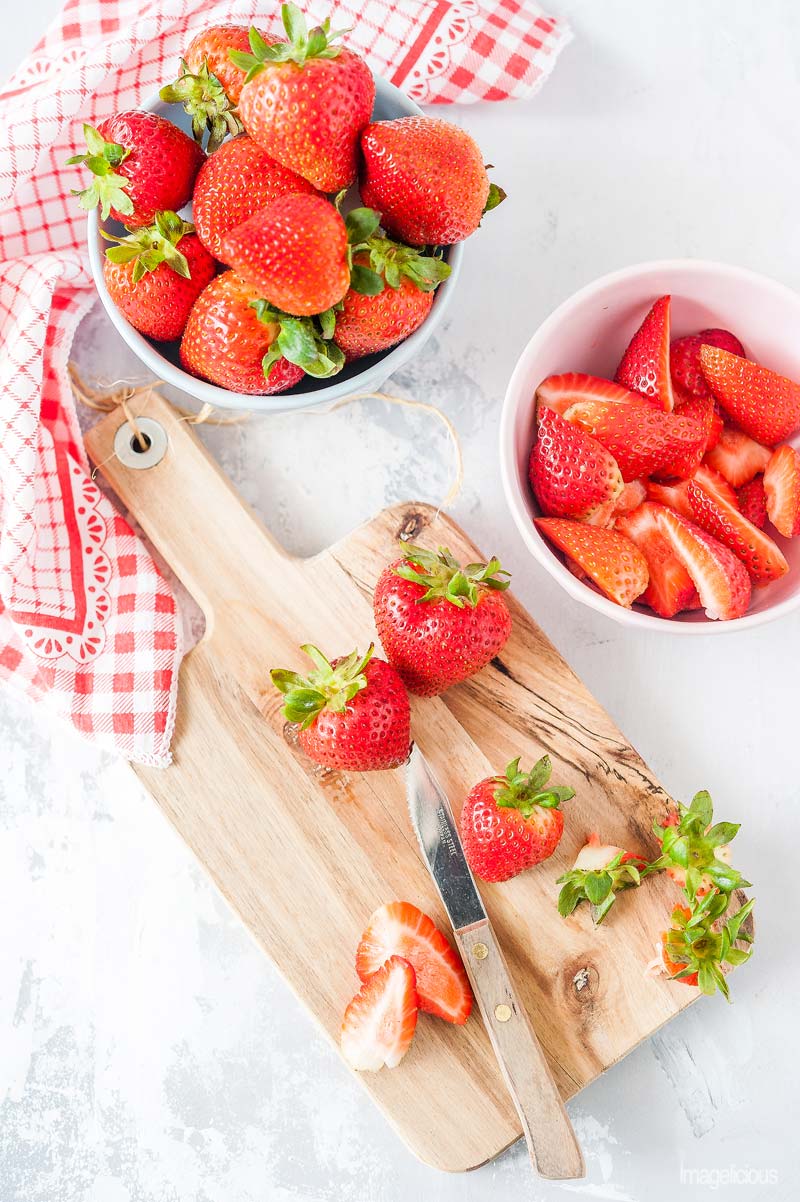 Top down view of a cutting board with a few strawberries being cut, another bowl with whole strawberries and a bowl with cut up strawberries are next to the cutting board