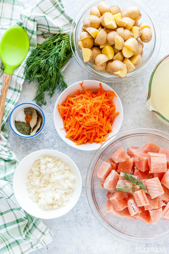 Top down view of all the ingredients that are needed to make Instant Pot Fish Soup. Bowls of diced onion, grated carrot, cubed salmon, cutup potatoes, seasonings, dill, and broth