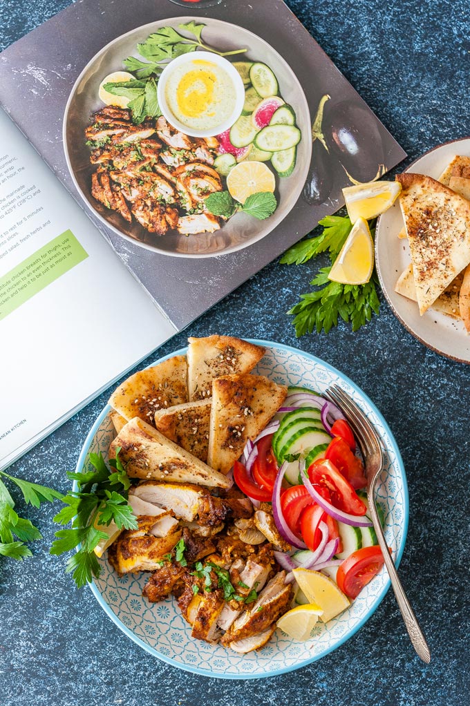 A bowl of Chicken Shawarma with some fresh tomatoes and cucumbers and pitas. More baked pitas on a plate next to the bowl. A cookbook with a photo of the same recipe is open and lying next to the food