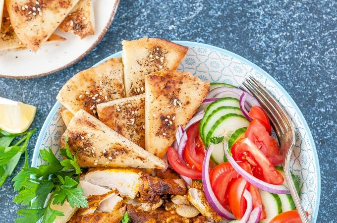 Top down view of a bowl of chicken shawarma with some fresh veggies and baked pitas. A plate of baked pitas is next to the bowl