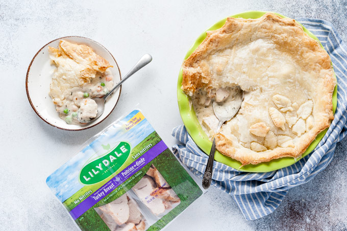 Top down view of a Whole Turkey Pot Pie with a slice cut out of it. A slice of Turkey pot pie is on a plate and a package of Lilydale turkey that is used in the recipe is also next to the plate