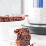 A stack of three Instant Pot Brownies with a cake plate with the rest of the brownies and an Instant Pot in the background