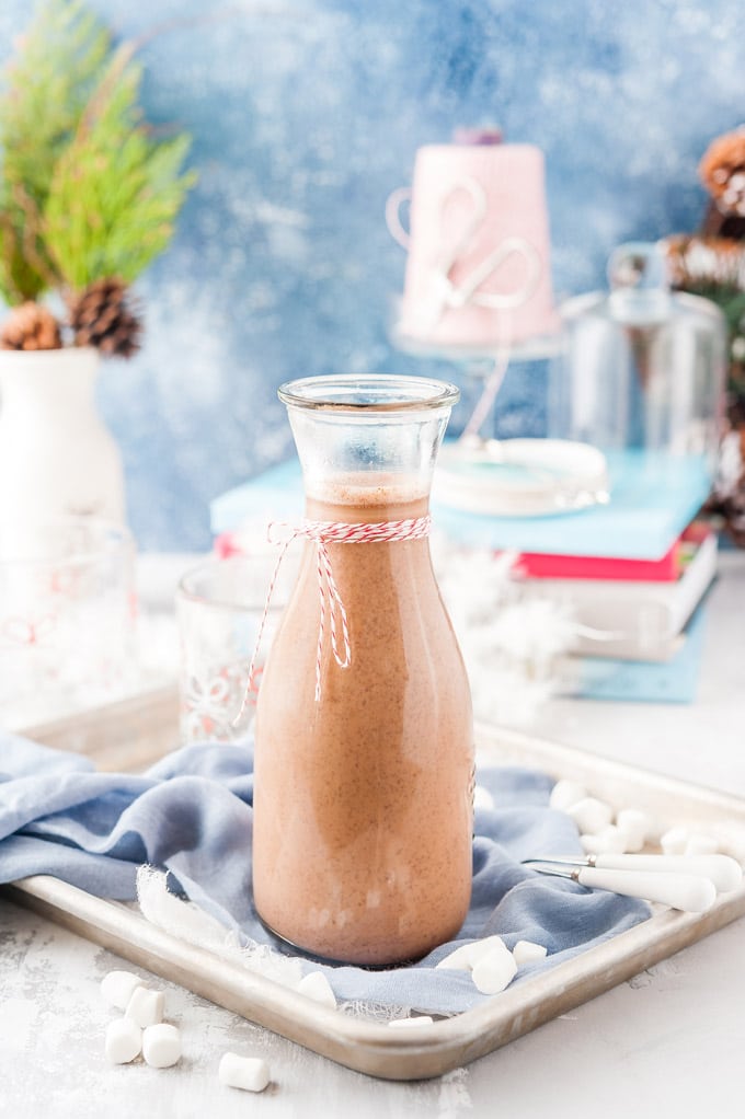 Tall carafe filled with Instant Pot Hot Chocolate on a tray with blue napkin and marshmallows around. More various Holiday themed accessories behind the hot chocolate
