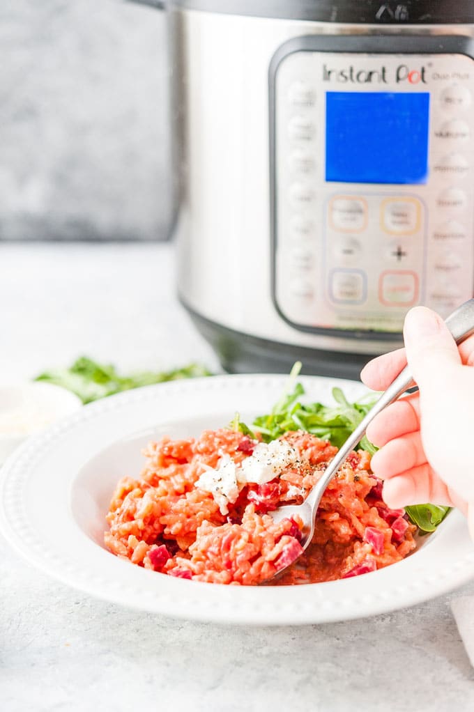 Bowl of Instant Pot Beet Risotto with a hand holding a spoon inside the bowl. Instant Pot is in the background