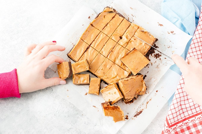 Butterscotch fudge on a piece of parchment paper with a hand taking one piece and another hand cutting a piece