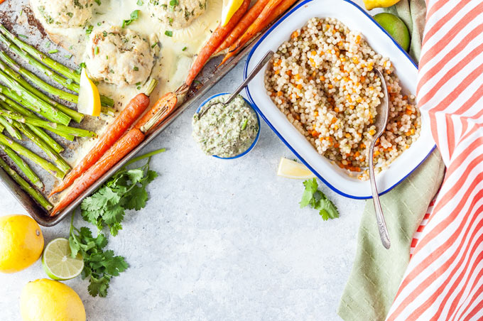 Top down view of a small part of sheet pan with vegetables and fish cakes, roasting pan with Israeli Couscous and a small bowl with carrot top pesto