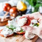 Hand holding a slice of tomato with goat cheese and dill spread. More tomatoes and herbs are in the background