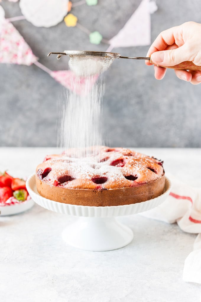Strawberry Cake on a cake stand with icing sugar falling over it.