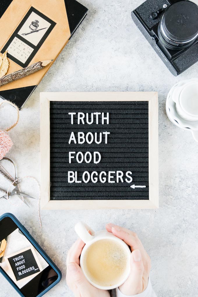 Cup of coffee, camera, notebook, letter board that says truth about food bloggers on a table.