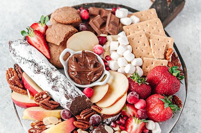 Top down view of a dessert board. It is filled with cookies, fruits, and chocolates.
